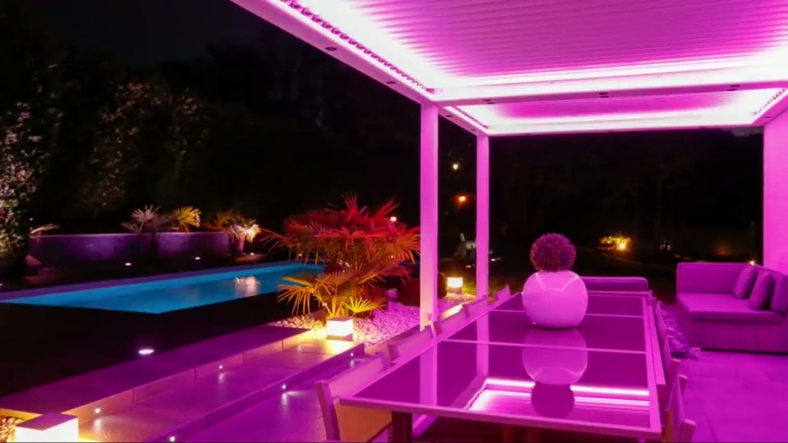 Havuz yanında pembe ışıkla aydınlatılmış pergola ve yemek masası